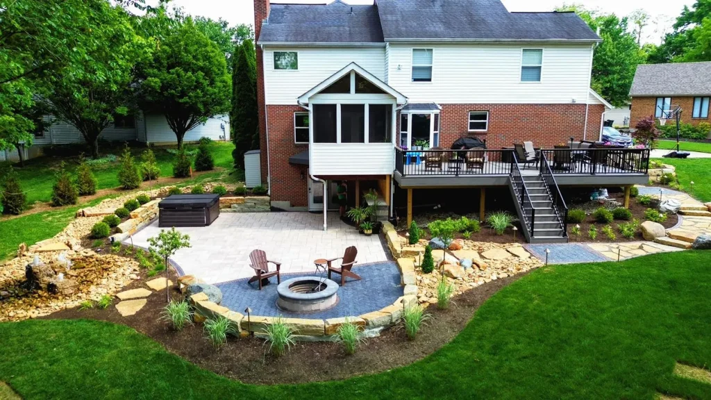 Aerial view of a backyard paver patio with a fire pit, deck, and stone retaining walls behind a brick home